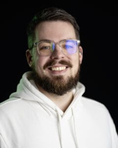 Mann mit Brille und Bart, der in einem weißen Kapuzenpullover lächelt.